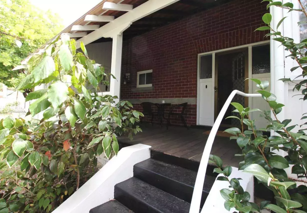 The front verandah of the Curtin family home with brown steps, a white railing and green leafy bushes. The house is brown with white doors.