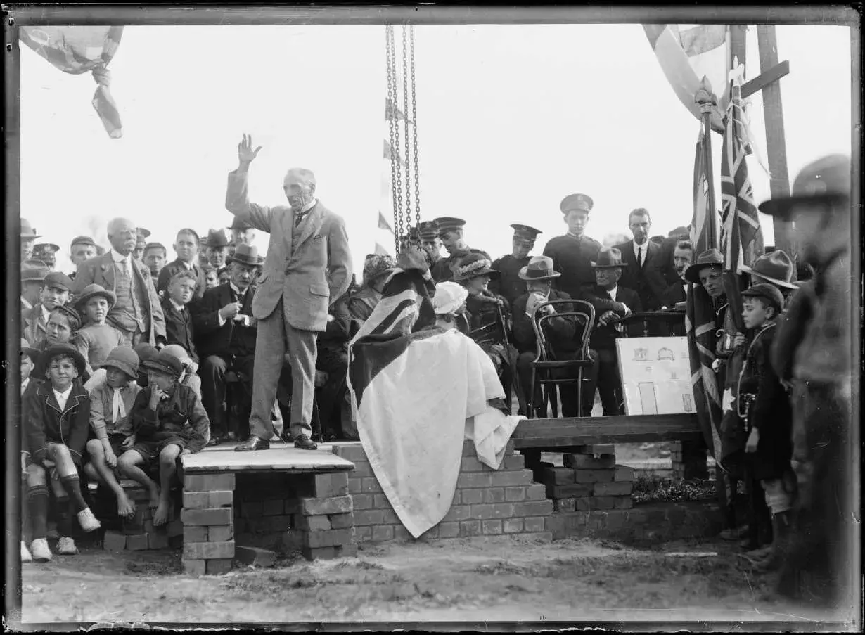 Billy Hughes stands on a stage surrounded by people while giving a speech and his hand is raised.  