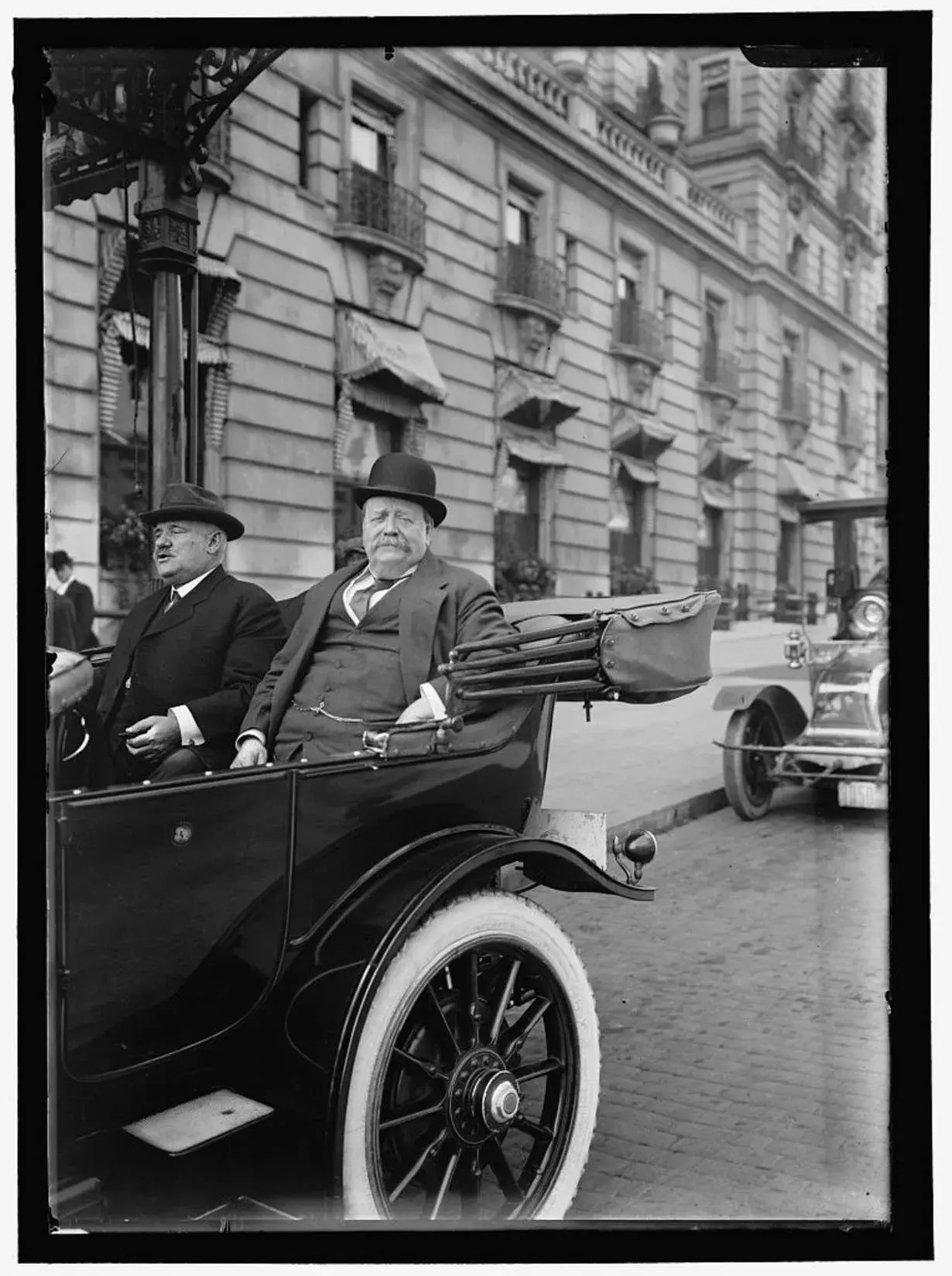 George Reid rides in the back of carriage with a top hat facing the camera.  