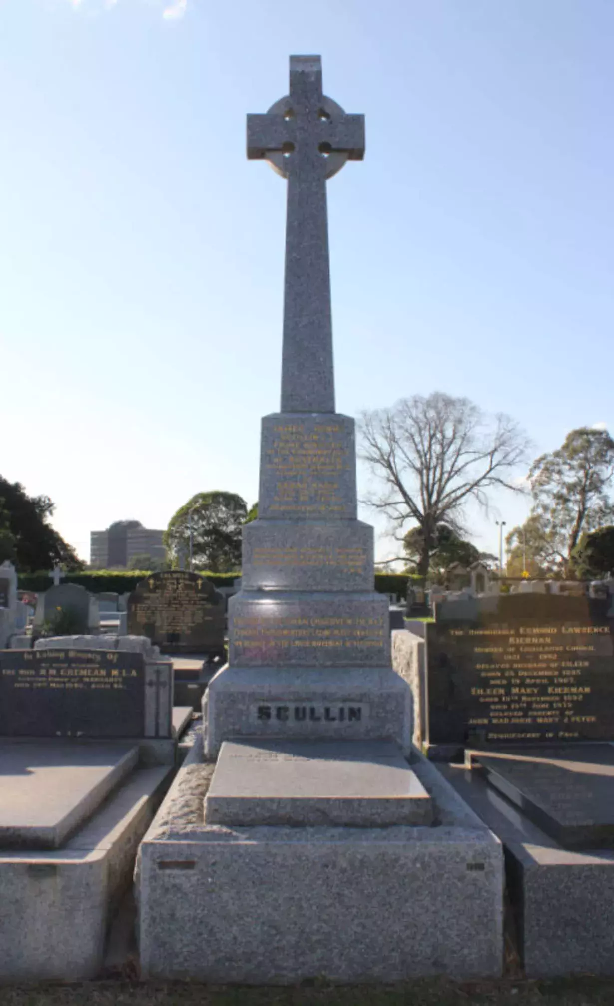 A stone monument in a cemetery with a square based and a tier of smaller stone layers leads to a a large cross. Words that can't be read are inscribed in gold. At the base of the monument is the engraved word 'Scullin'. 