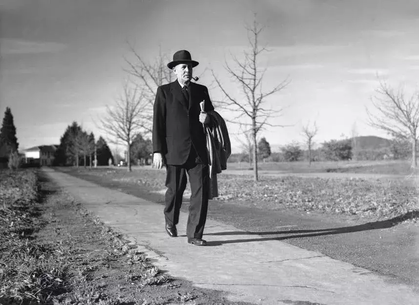 Ben Chifley walking with a pipe in his mouth and a coat over his arm.  