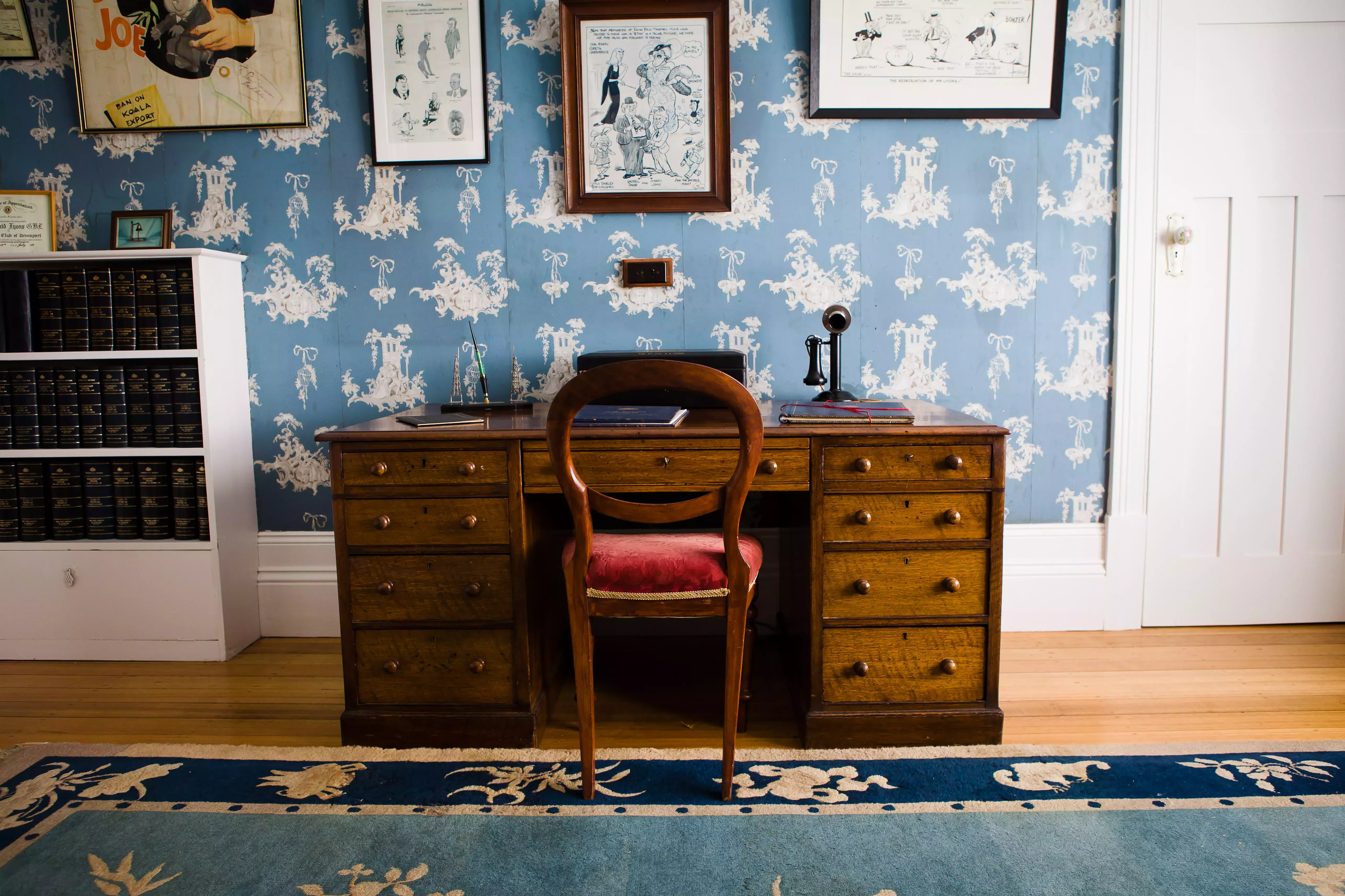 A brown desk with a red chair in a study with blue wallpaper.  