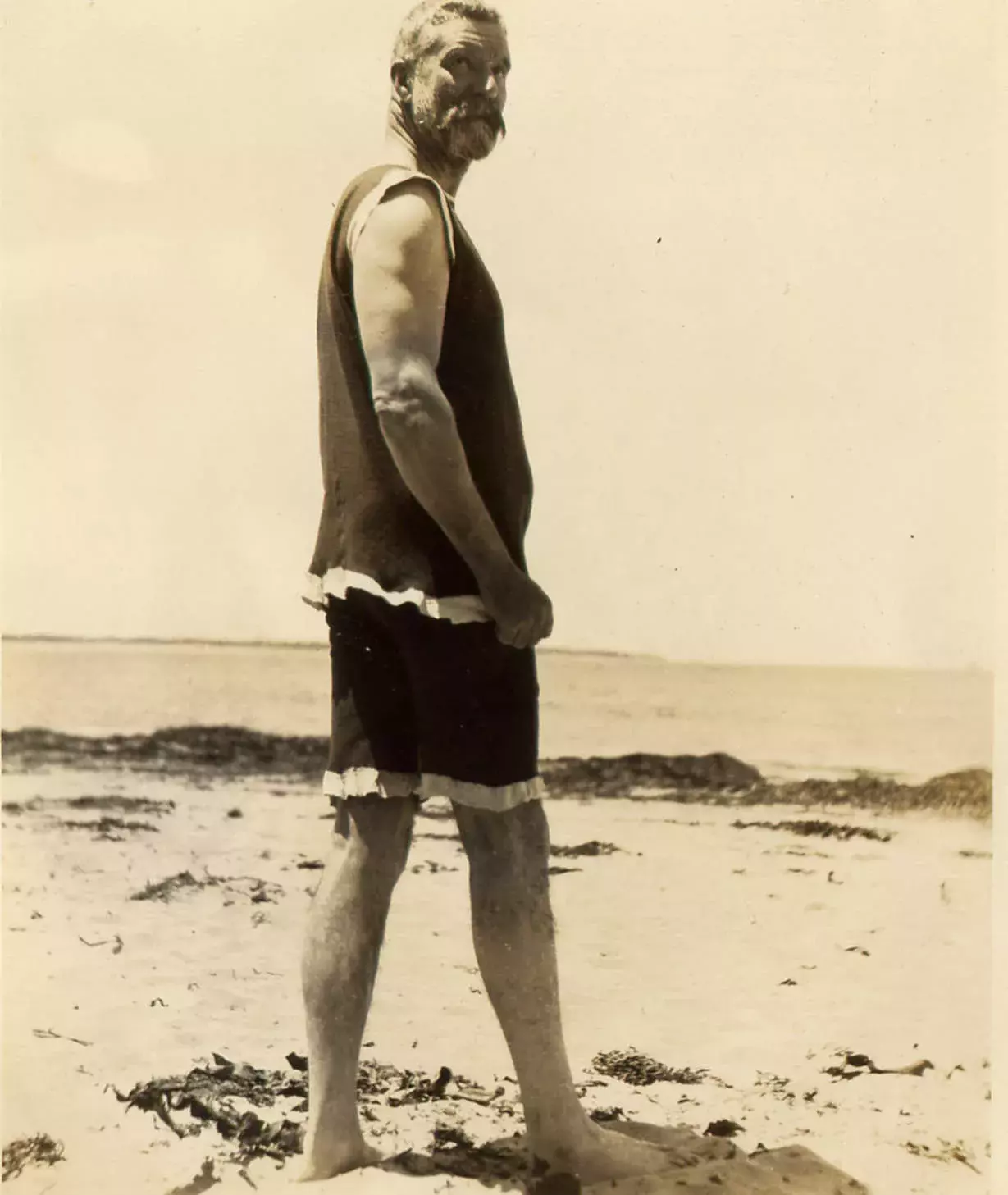 Alfred Deakin wearing a singlet and bathing shorts standing on the beach looking back at the camera.