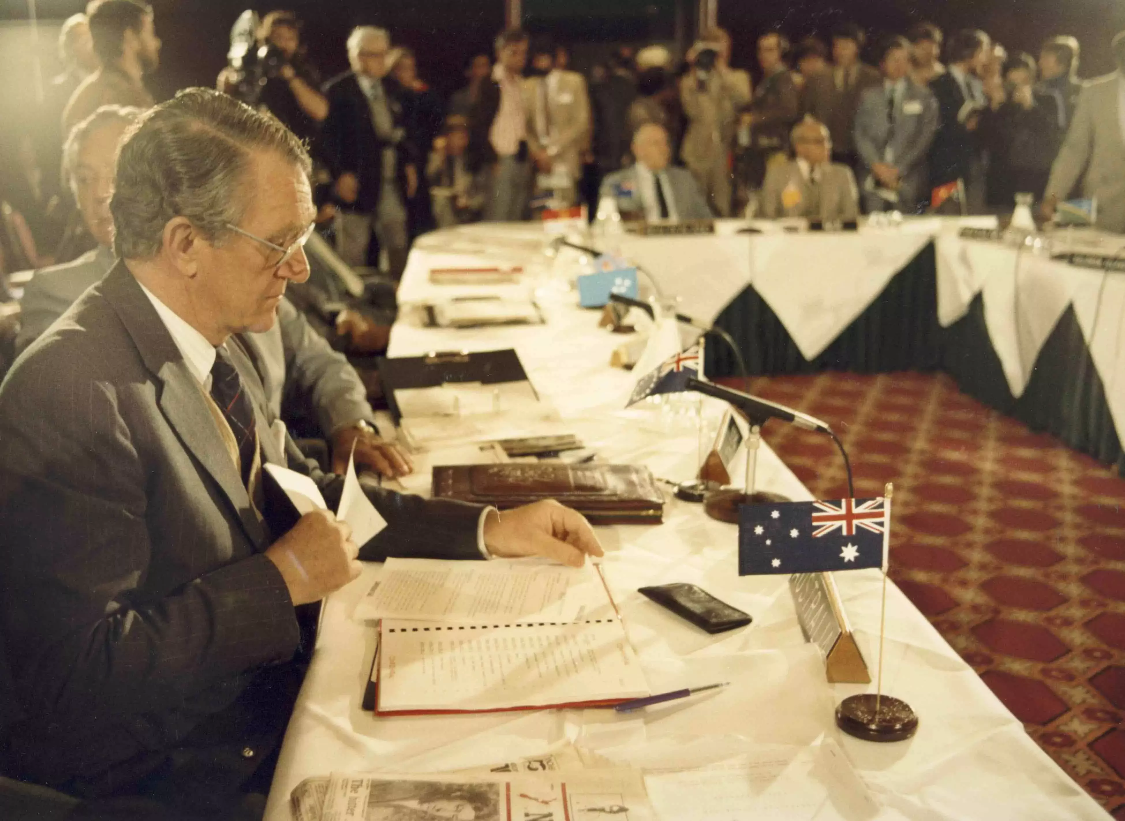 Malcolm Fraser sits at the end of a long U-Shaped table with a pile of papers in front of him that he reads. There is a small Australian flag on the table. The entire table is filled with people also looking at documents.  